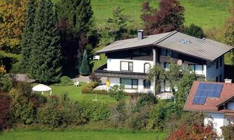 A modern house surrounded by green meadows and trees. In the garden, there is a lounge chair, and the sun is shining.
