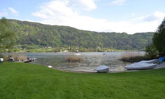 A quiet lake surrounded by green mountains. In the foreground, boats and a meadow can be seen.