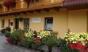 A cozy house with balconies adorned with red and green plants. Flower beds in vibrant colors decorate the entrance area.