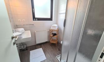 A modern bathroom with a shower, sink, and towels. The room is bright and clean, with tiled flooring and a window.