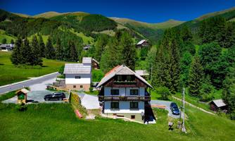 A picturesque house in nature, surrounded by green hills and forests. The road and some cars are visible nearby.