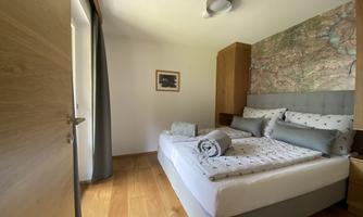 A modern bedroom with a double bed and light wooden furniture. The walls are decorated with a world map and large windows let in plenty of light.