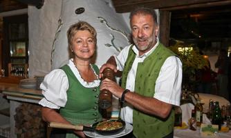 A couple in traditional clothing stands in a restaurant. They are holding a pan with food and a pepper mill while smiling at the camera.
