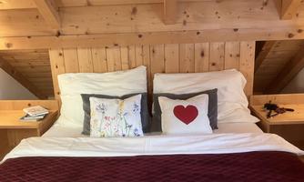 A cozy bed under a wooden ceiling. The pillows and blanket are in soft colors, with a heart-shaped pillow as a centerpiece.