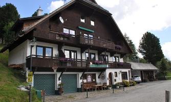 A traditional two-story wooden house with balconies and flowers. In front of the building are garden furniture and a quiet street.