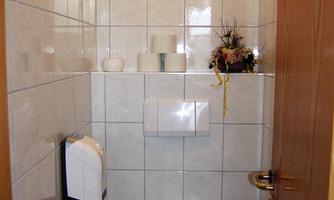 A small bathroom with white tiles and a toilet. On the wall, there are toilet paper holders and some decorative elements.