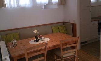 A cozy dining area with a wooden table and chairs. The walls are light, and there are green cushions on the bench.