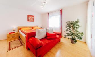 A cozy bedroom with a large bed and orange pillows. Additionally, there is a red sofa and a plant in the room.