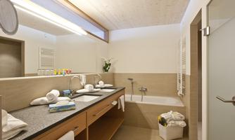 A modern bathroom with two sinks, a bathtub, and large mirrors. Towels and toiletries are visible on the shelf.