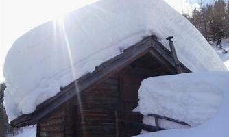 A small wooden house covered by a thick layer of snow. The snow hangs over the roof and it seems to be a lot of winter.