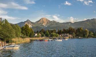A tranquil lake with boats and wooded shores. In the background, impressive mountains rise under a blue sky.
