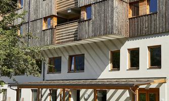 A modern building with a wooden facade and large windows. In front of the house is a terrace with seating.