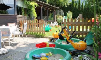 An inviting garden with toys for children, including a sandbox and a digger. In the background stands a cozy little house with a terrace.