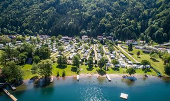 A beautiful campsite by the shore of a lake, surrounded by green trees and mountains. Many motorhomes and tents are scattered across the meadow.
