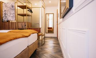 A modern bedroom with wooden flooring and a large bed. In the background, a mirror and a wardrobe are visible.