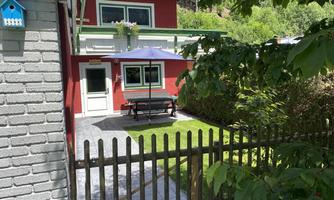 A red house with a small garden and a seating area under a sunshade. A wooden plank fence surrounds the green lawn.