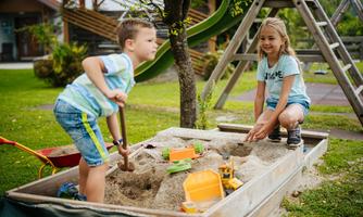 Two children are playing in the sandbox. A boy is digging with a shovel while a girl is playing with toy cars.