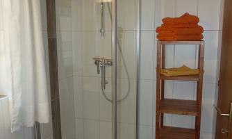 A modern shower with glass swinging doors and a showerhead. Orange towels are placed on a wooden shelf.