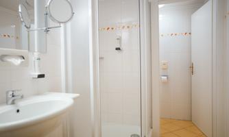 A bright bathroom with a shower and a sink. The walls are lined with white tiles, and there is a mirror as well as a door to another room.