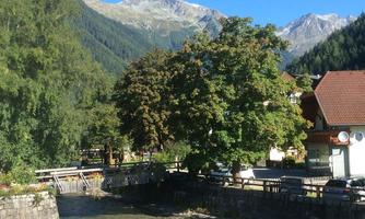 A picturesque valley with mountains in the background and green foliage. In the foreground, a clear stream flows beside a wooden bridge.