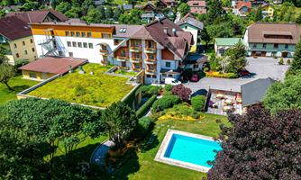A picturesque hotel complex with a green roof, surrounded by trees and gardens. In the foreground, there is an inviting pool.
