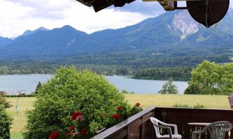 A beautiful view from a balcony overlooking a lake and the surrounding mountains. Green trees and red flowers adorn the foreground.