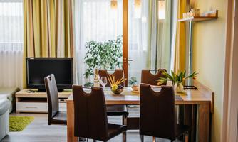A modern dining room with a wooden table and four chairs. In the background, there is a television and plants visible, the light is bright and welcoming.