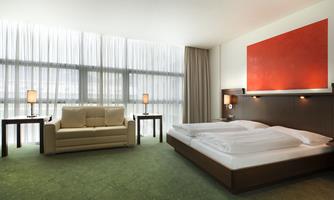 A modern hotel room with a double bed and a comfortable sofa. Large windows with curtains let in plenty of light.