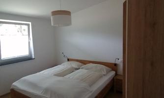 A bright bedroom with a wooden double bed. The walls are plain white, and a window lets in plenty of light.
