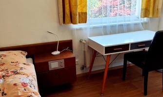 A cozy room with a single bed, a nightstand, and a desk. The yellow curtains let in plenty of daylight.