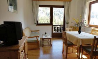 A cozy living room with a wooden chest of drawers, a table, and chairs. Larger windows offer a view of nature.