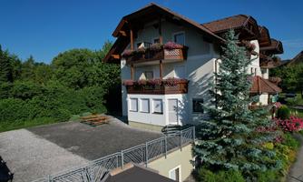 A large two-story house with balconies and a well-maintained garden. The sky is clear and there is a parking space as well as an outdoor seating area.