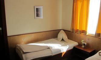 A simple room with two beds and a window framed by yellow curtains. A nightstand with a vase and a framed picture can also be seen.