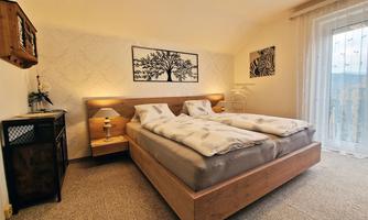 A cozy bedroom with a double bed and modern wood design. Artwork hangs on the wall, and a window provides ample daylight.