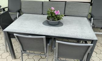 A modern table with a gray surface and a flower pot in the center stands on a paved floor. Four chairs with gray seat cushions are arranged around the table.