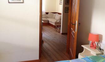 Insight into a bedroom with a bed and a nightstand. Through the open door, a living area with a sofa can be seen.
