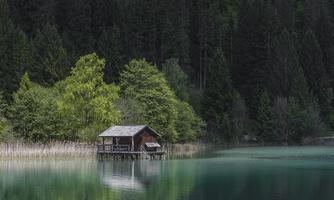 A cozy cabin sits by the shore of a calm, clear lake. Surrounded by lush greenery and tall trees, the cabin reflects in the water.
