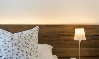A cozy bedroom with a wooden bed and pretty bedding. Pictures of picturesque landscapes hang on the wall, and a nightstand with a lamp stands next to the bed.