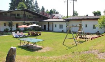 A cozy garden with lawn, a table tennis table, and a playground. In the background, there is a modern accommodation with sun umbrellas and loungers.