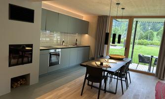 A modern kitchen with sleek gray cabinets and a dining table. Through large windows, one looks out onto a green landscape.