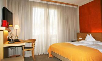 A modern hotel room with a large window and light curtains. The room is simply furnished, with a bed, a desk, and a cozy chair.