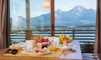 A lavish breakfast table with fresh rolls, fruit, and beverages. In the background, mountains and a lake can be seen.