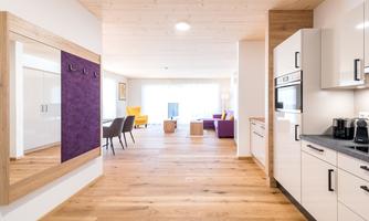 A modern, bright living space with wooden floors. The open kitchen leads to an inviting living area with colorful furniture.