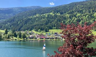 A peaceful lake with a small sailboat and green hills in the background. The shore is surrounded by trees and picturesque houses.