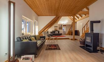 A modern living room with wooden floors and a wooden ceiling. There is a black sofa, a fireplace, and light-colored furniture.