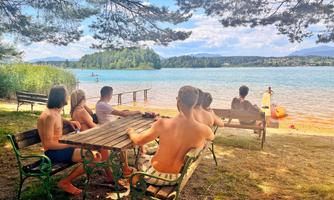 A group of people is sitting at a table by the water and enjoying the view. In the background, the lake and a picturesque landscape can be seen.