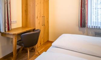 A simple bedroom with two beds, a wooden desk, and a practical wardrobe. The walls are bright, and the light is friendly and inviting.