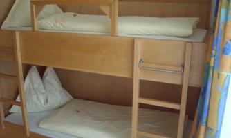 A children's bed with two levels made of light wood. The mattresses are equipped with light-colored bedcovers and pillows.
