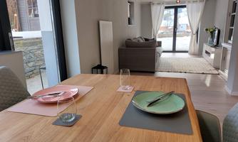 A modern dining room with a wooden table set with colorful dishes. In the background, there are large windows that let in plenty of light, and a cozy living area.