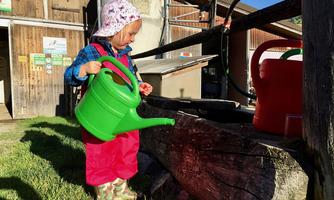 A small child is watering with a green watering can. The background shows a wooden barn and sunny weather.